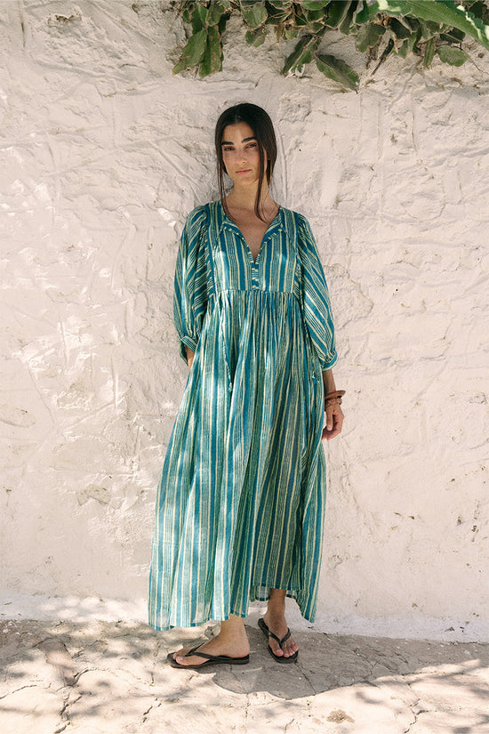 Full-length front view of woman wearing the Daughters of India Kyra Maxi Dress in Spearmint against a white wall in Greece, showing the flowing green striped silhouette