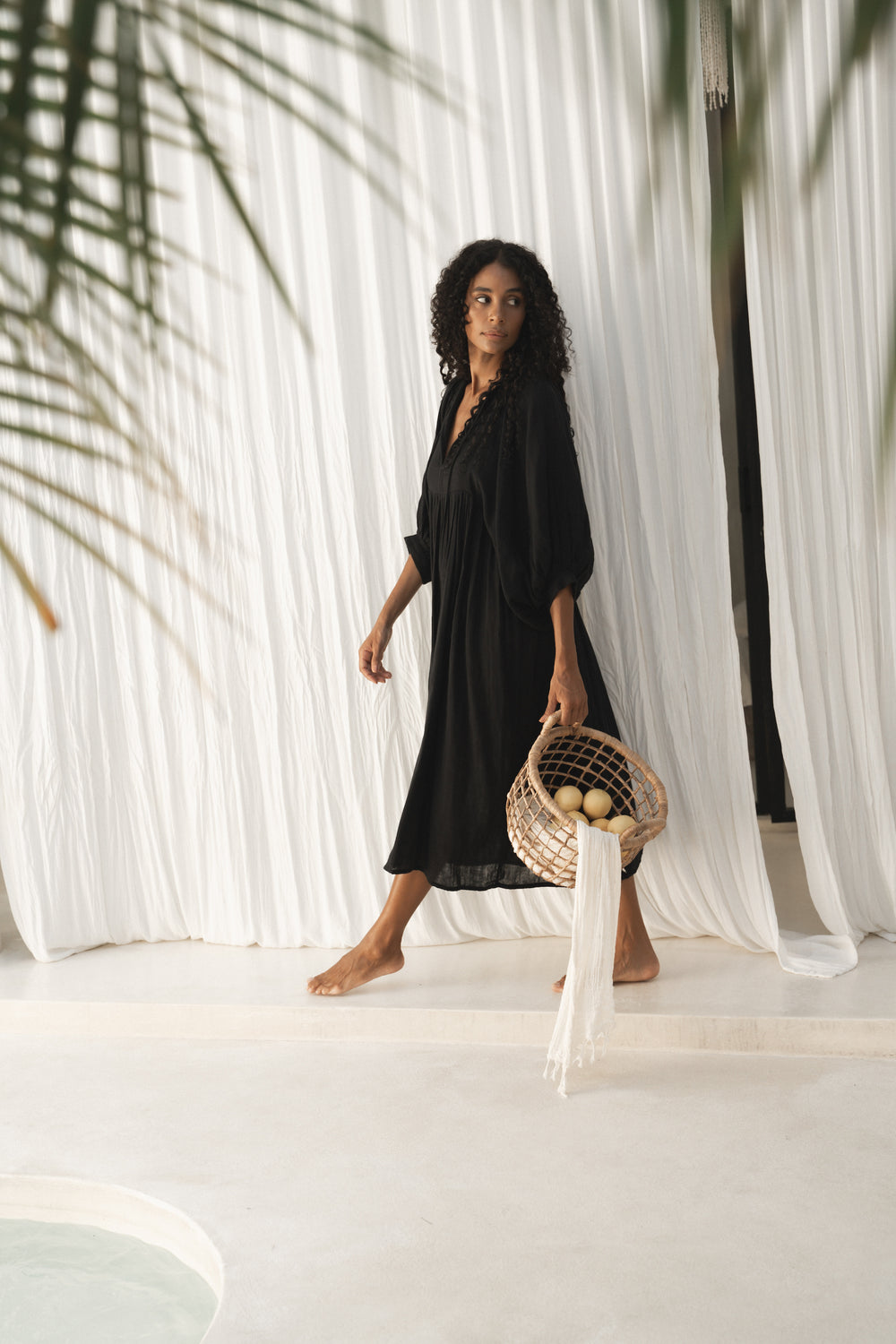 Daughters of India Kyra Midi Dress in Black Gauze — walking with woven basket by white drapes, palm shadow